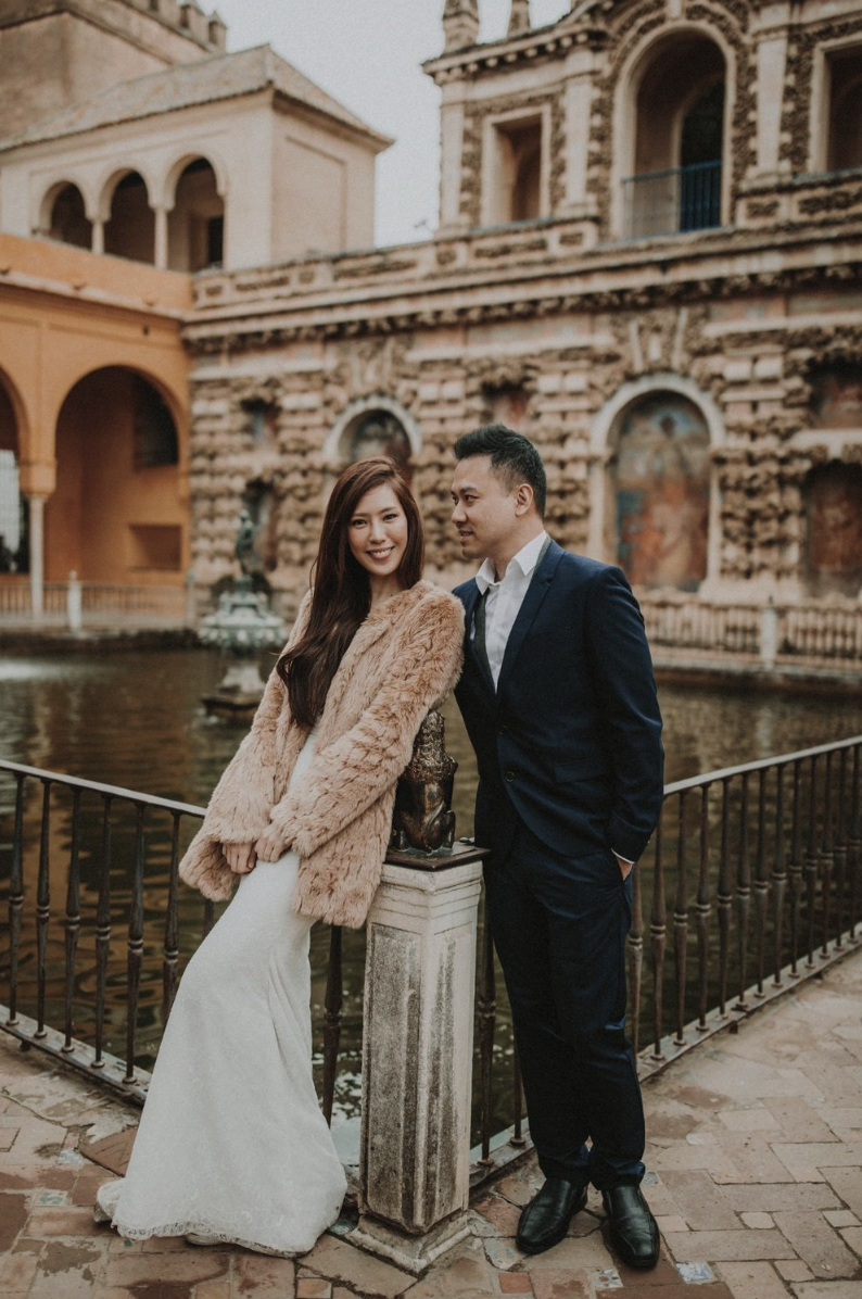 A couple standing near a railing by a pond with an ornate, historic building in the background. The woman is wearing a white dress and a furry coat, while the man is dressed in a navy suit with a white shirt.