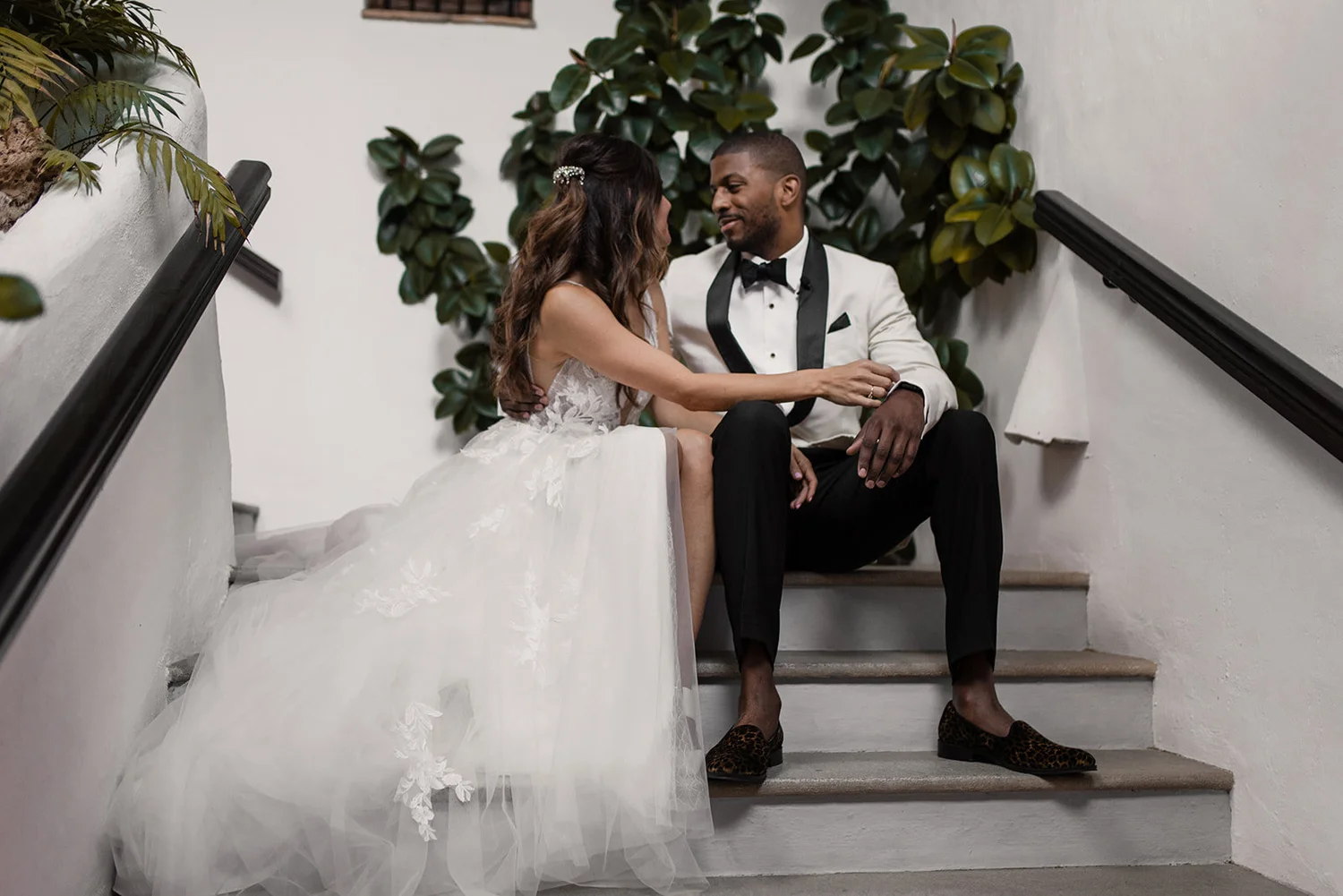 A bride and groom sitting on stairs, facing each other, sharing a moment. The bride is wearing a white wedding gown and the groom is dressed in a tuxedo with leopard print shoes. They are indoors with greenery in the background.