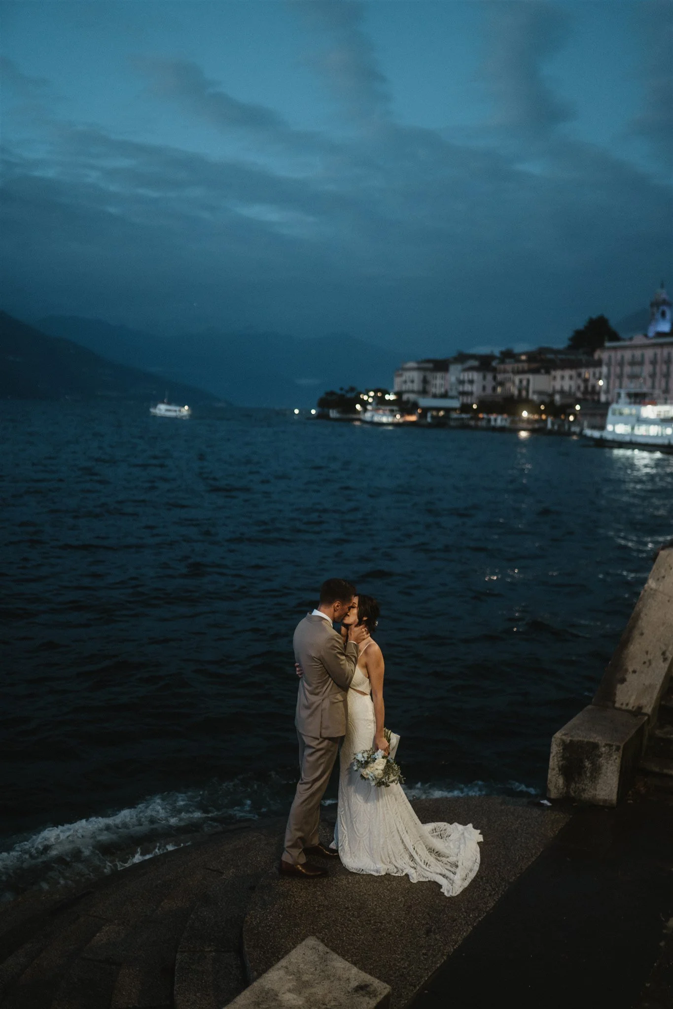 A couple dressed in wedding attire sharing an intimate moment by the water at dusk, with mountains and buildings in the background.