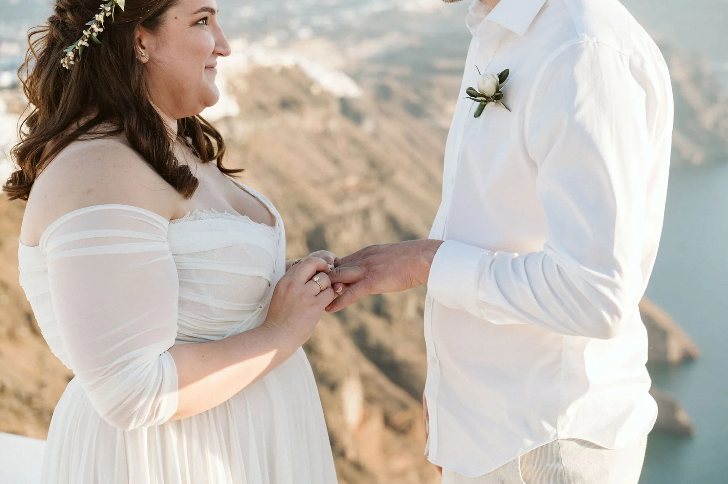 A bride and groom holding hands during a wedding ceremony outdoors with a scenic view of cliffs and water in the background.