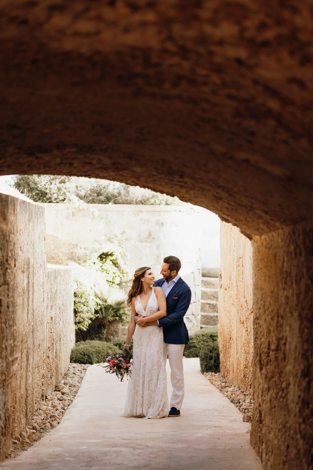 A bride and groom standing together on a pathway under a stone archway, smiling and looking into each other's eyes, with the bride holding a bouquet of flowers.