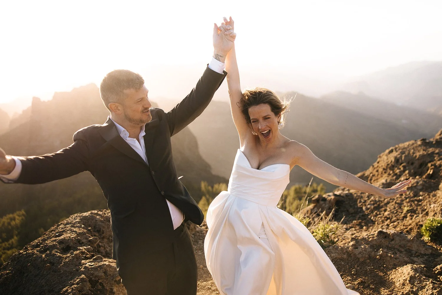 A couple in wedding attire celebrating outdoors, with the man holding the woman's hand high, both smiling and joyful against a mountainous sunset background.