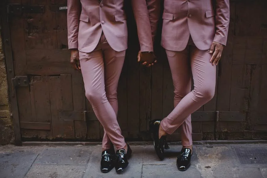 Two people dressed in pink suits holding hands, standing against a wooden door.