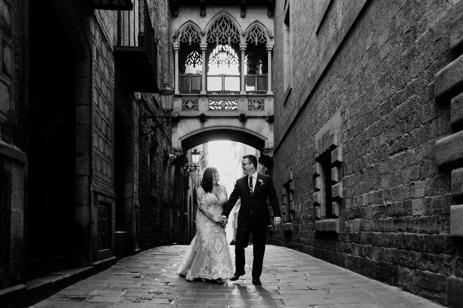 A couple in wedding attire walking hand in hand in a narrow cobblestone alley with gothic architecture and ornate windows above.