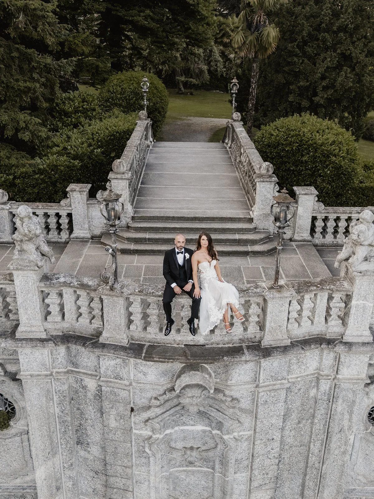 A newlywed couple dressed in wedding attire sitting on a stone balcony, with a staircase and lush greenery in the background.