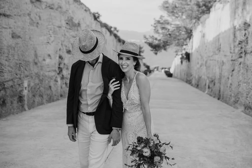 A woman in a wedding dress holding a bouquet and smiling, walking with a man in a blazer, white pants, and a hat, in a scenic outdoor area with rocky walls and trees.