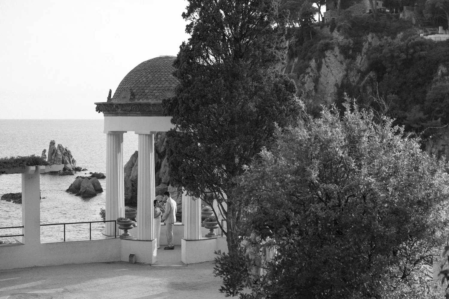 A black and white photo of a man and woman standing together under a small classical pavilion with columns, near the water, with a hillside covered in trees and houses in the background.