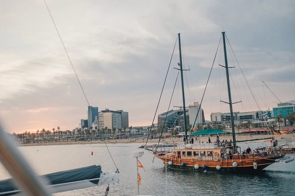 View of a marina with sailboats docked and a city shoreline with modern buildings in the background during sunset.