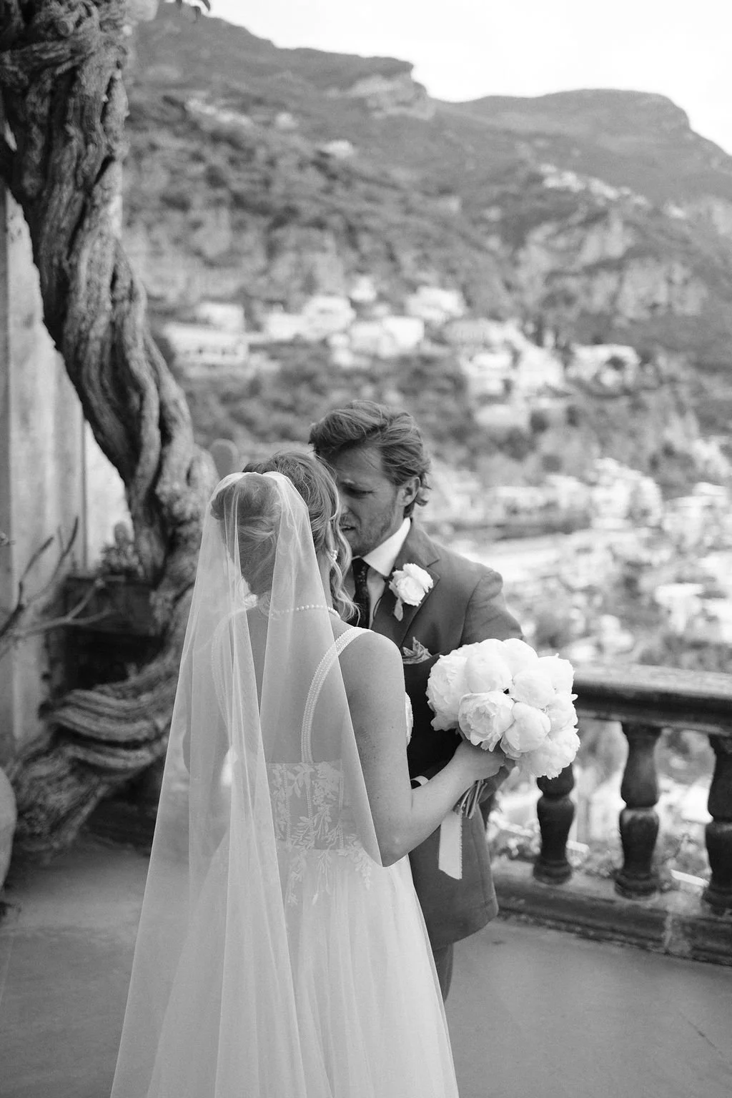 Bride and groom sharing a moment on a balcony with scenic mountains in the background, groom holding a bouquet of flowers, black and white photo.