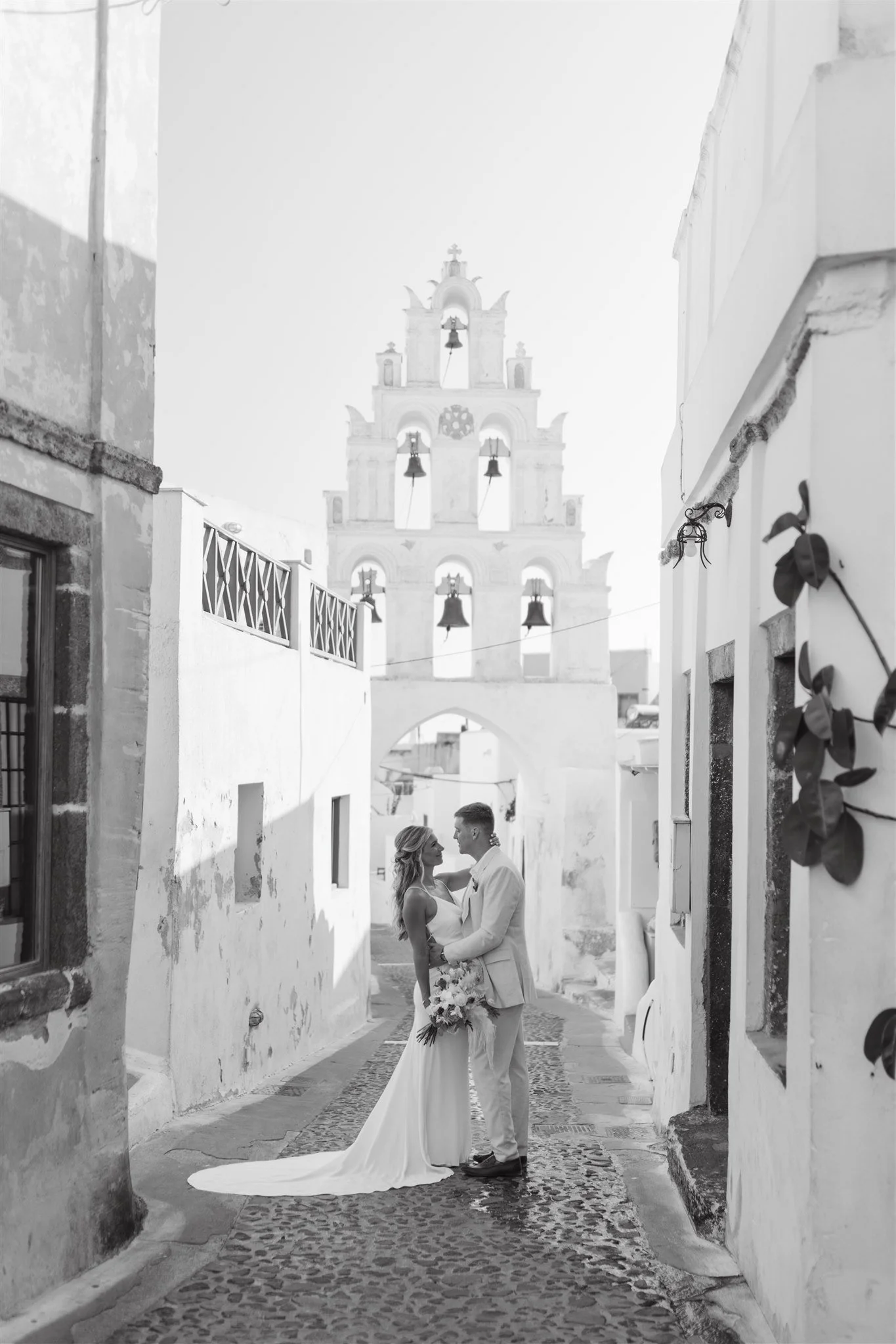 A bride and groom standing close together on a cobblestone street in a historic European town, with a white church with multiple bells and architectural details in the background.