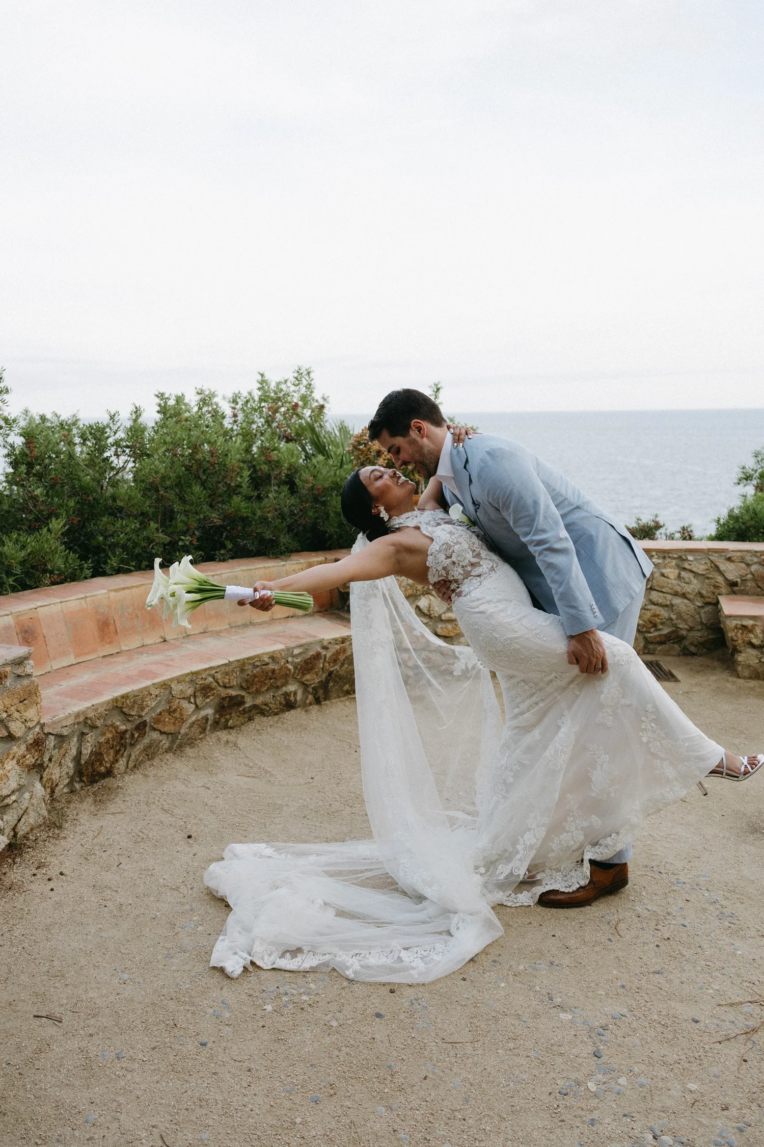 A bride and groom are posed in a romantic dance scene outdoors near the ocean under an overcast sky. The bride is wearing a white lace wedding gown and holding a bouquet of white calla lilies, and the groom is dressed in a light blue suit. He is dipp
