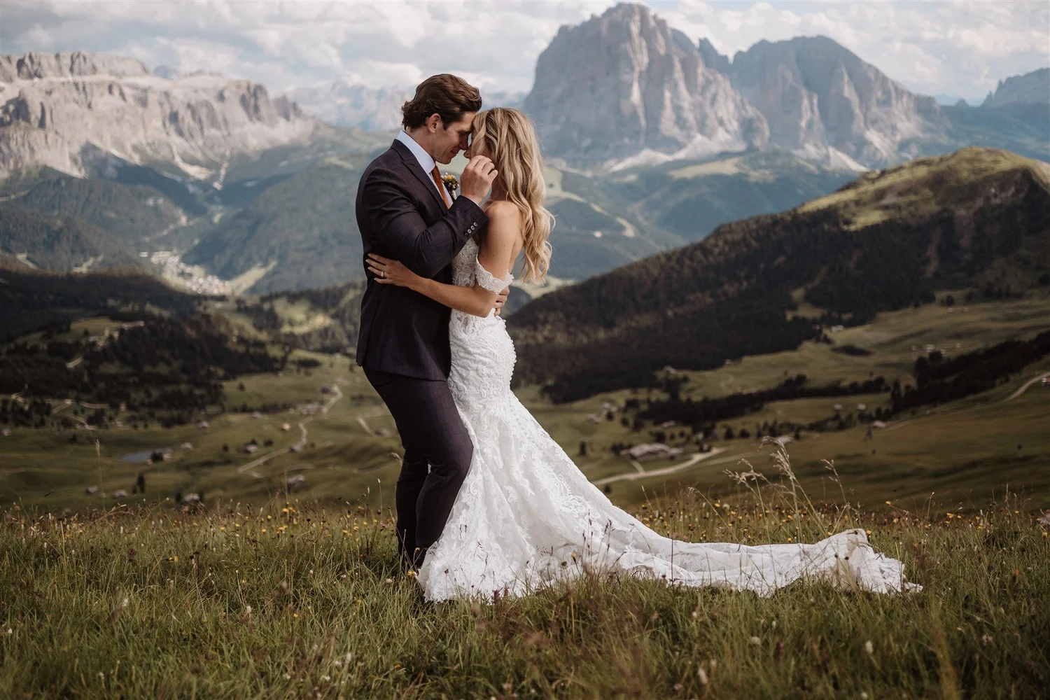 A bride and groom embrace in a mountain meadow with rolling hills and towering mountain peaks in the background. The bride is wearing a white lace wedding gown, and the groom is in a dark suit with a white shirt and tie. They are facing each other wi