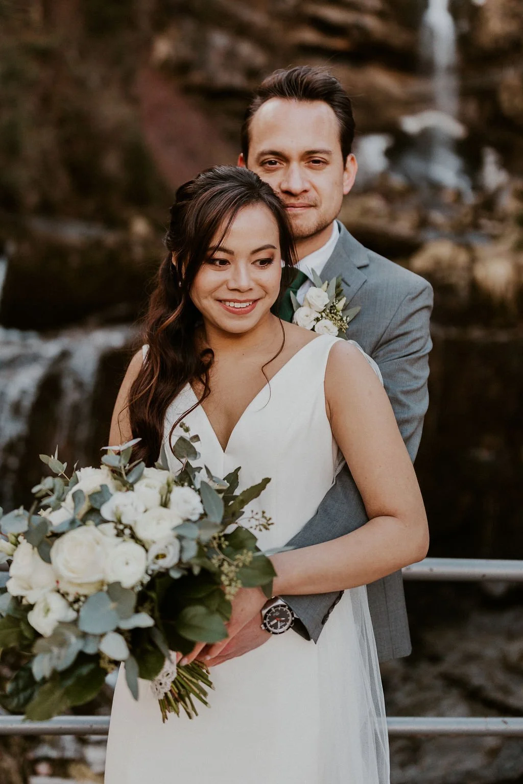 A newlywed couple embracing outdoors by a rocky waterfall. The bride is holding a bouquet of white flowers with greenery, and the groom is standing behind her in a gray suit with a white shirt and boutonniere. Both are smiling softly, looking content
