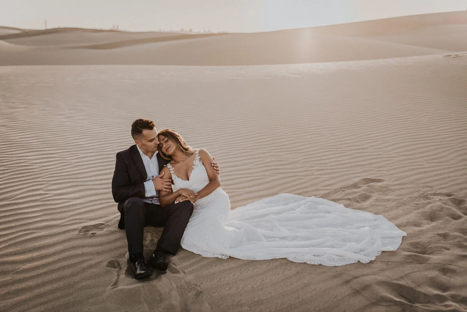 A couple in wedding attire sitting on sand dunes in a desert, with the groom dressed in a black suit and the bride in a white wedding dress, embracing and enjoying the moment.