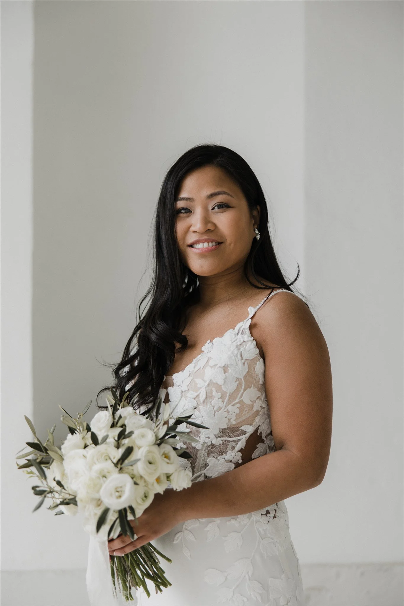 A smiling bride with long dark hair wearing a white lace wedding dress and holding a bouquet of white roses and greenery against a plain white wall.