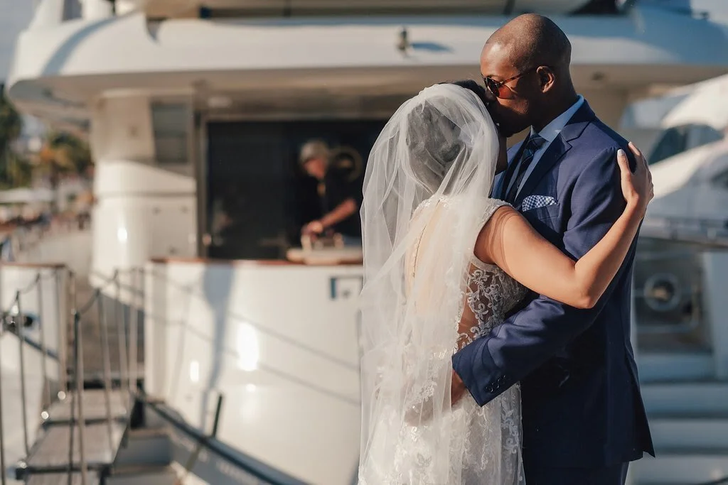 A bride and groom sharing a kiss on a yacht during sunset. The bride is wearing a veil and lace wedding dress, and the groom is in a navy suit with sunglasses.