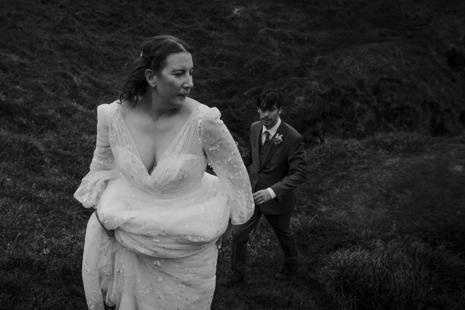 A black and white photo of a woman in a wedding dress walking outdoors with a man in a suit in the background.