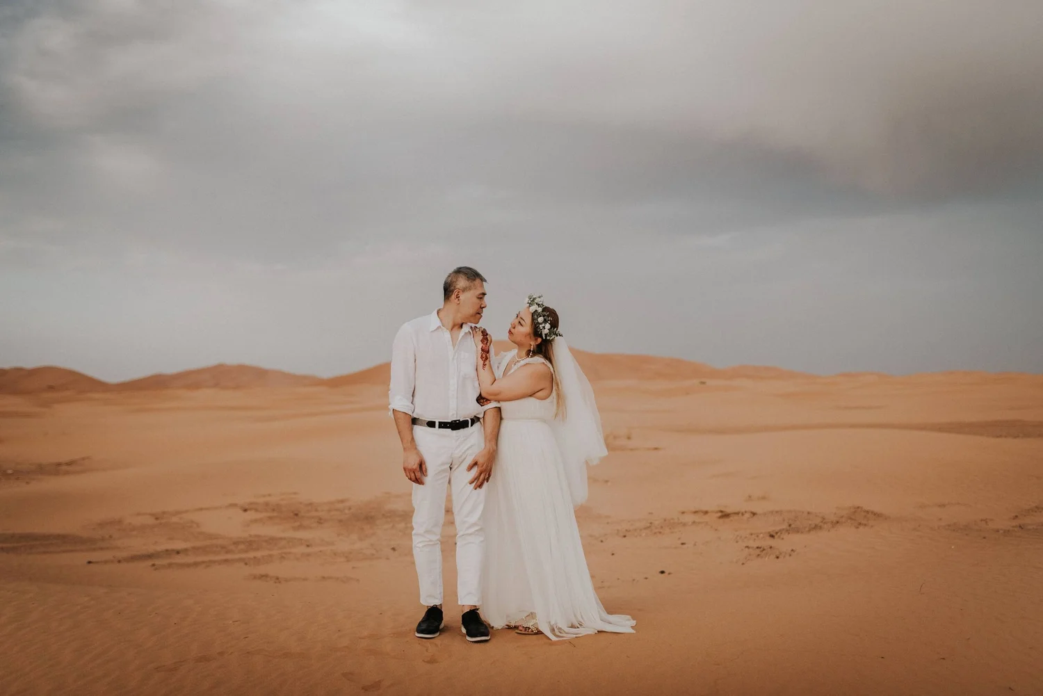 A couple in wedding attire standing in a desert with sand dunes in the background, under a cloudy sky.