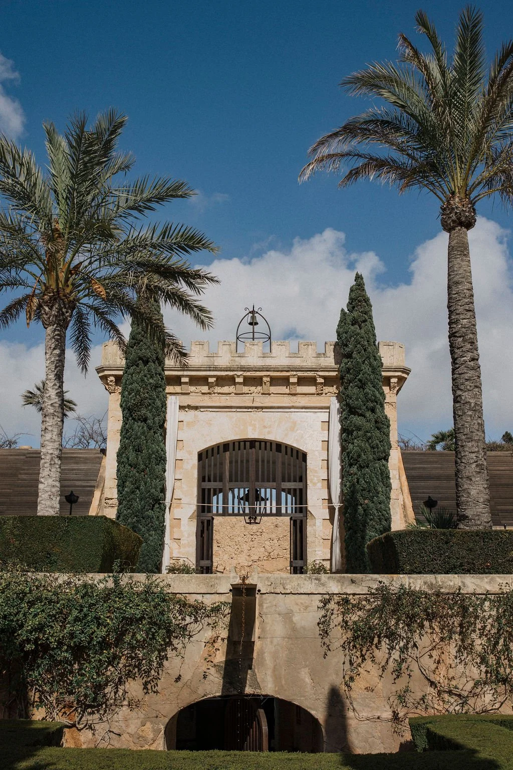 A historic stone building with a crenellated roof, flanked by two tall cypress trees and two palm trees, under a partly cloudy blue sky.