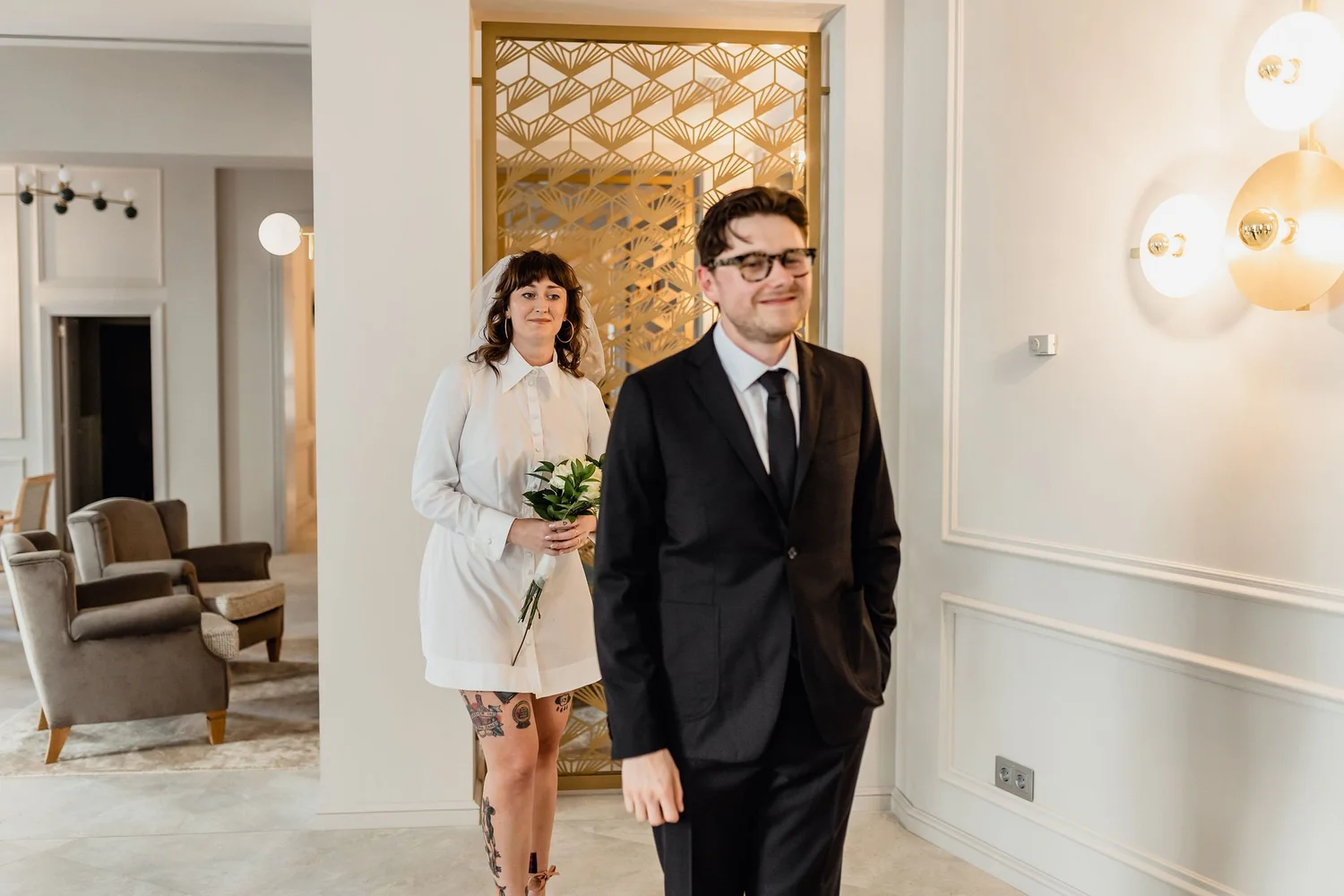 A couple dressed in wedding attire appears to be at a wedding or special event in a well-lit room with white walls and elegant decor. The woman is holding a bouquet and wearing a white dress with tattoos visible on her legs. The man is wearing a blac