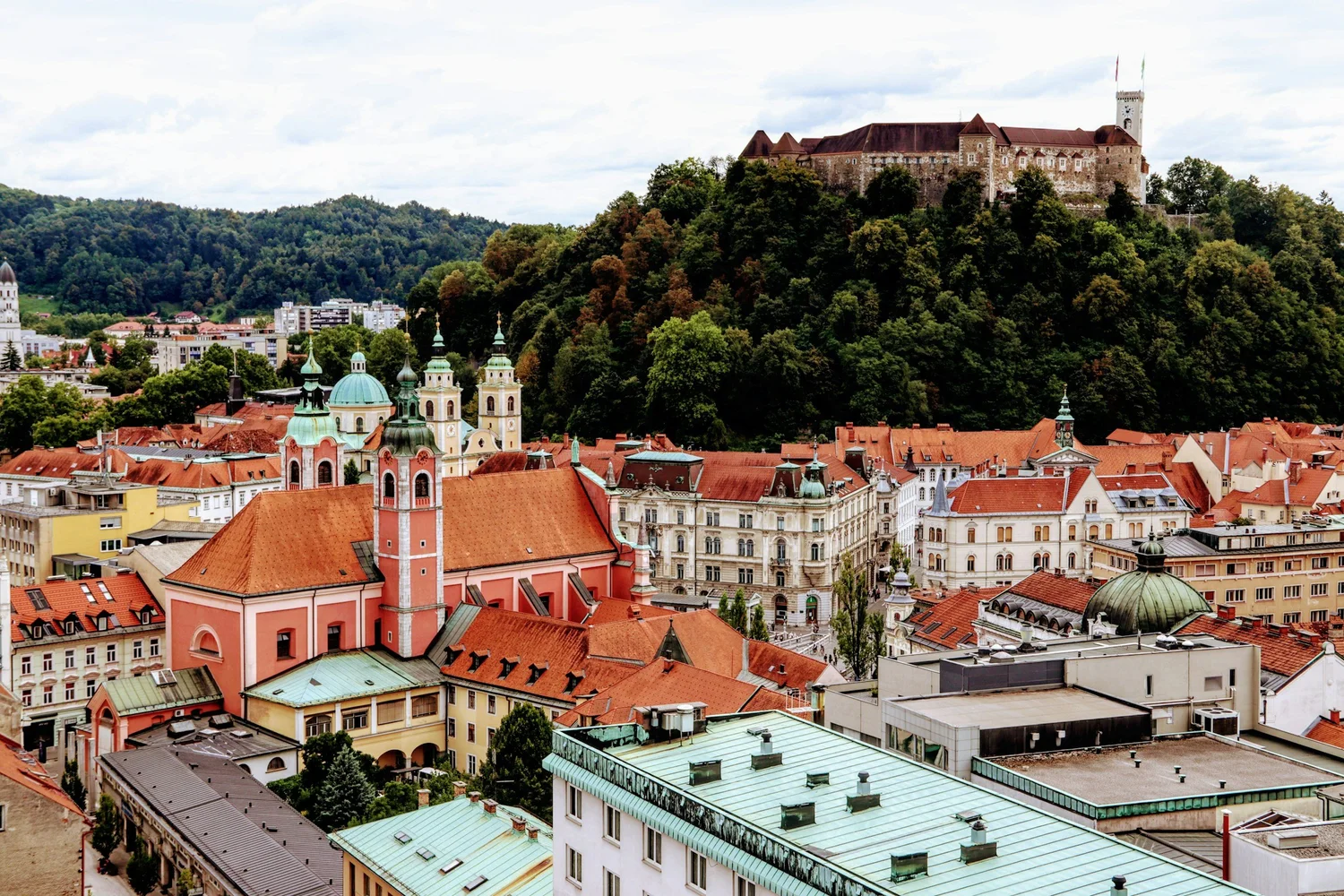 A view of a European city with historic buildings, domed churches, red-tiled roofs, and a castle on a hill covered in trees under a partly cloudy sky.