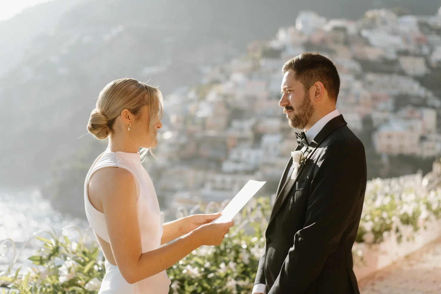 A bride and groom exchanging wedding vows outdoors with scenic hillside buildings in the background.