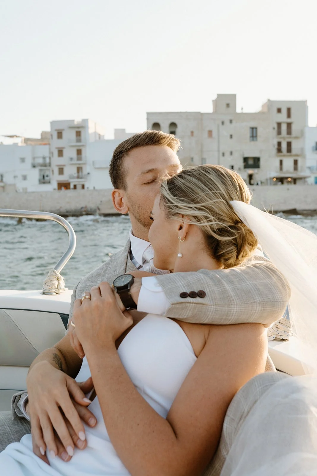 A newlywed couple kissing on a boat near the water with a backdrop of white buildings.
