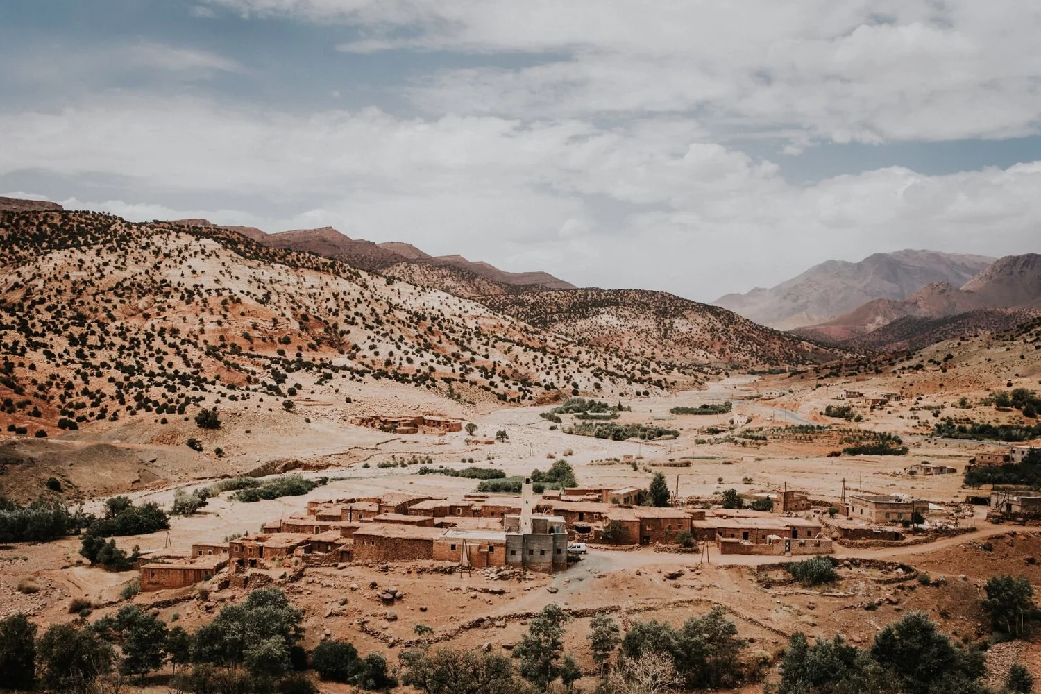 A small rural village in a dry, desert landscape with mountains in the background. The village consists of several adobe-style buildings surrounded by sparse trees and shrubs.