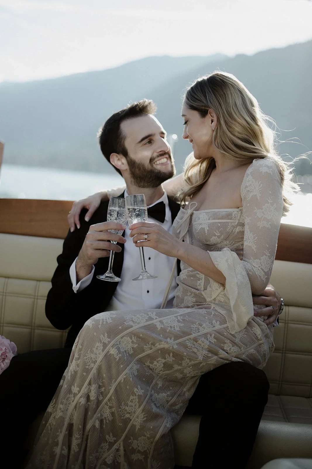 A newlywed couple celebrating with champagne on a boat with mountains and water in the background.