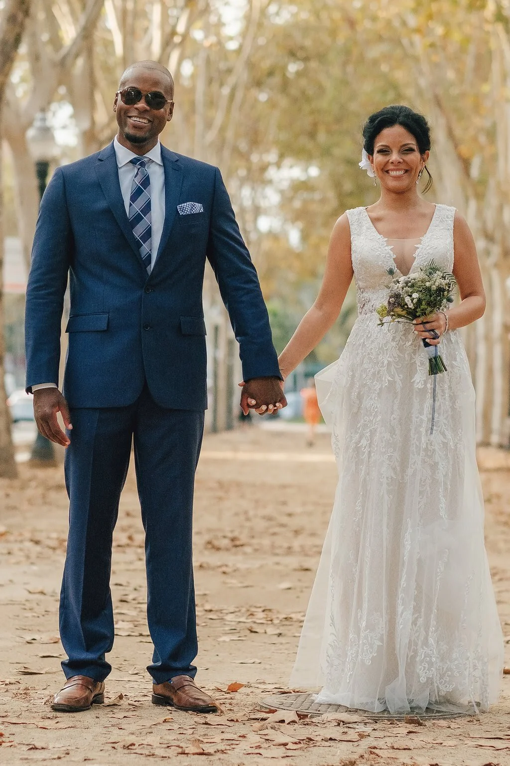 A couple dressed in wedding attire holding hands, walking outdoors on a leaf-covered pathway lined with trees. The man is wearing sunglasses and a navy blue suit with a tie, while the woman is holding a bouquet, wearing a lace wedding gown and smilin