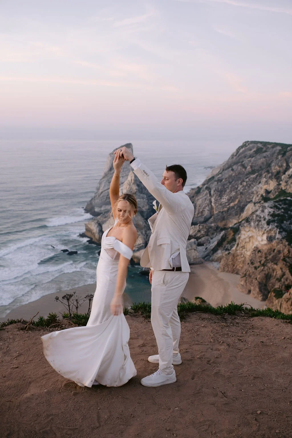 A newlywed couple dancing on a cliffside overlooking the ocean during sunset.