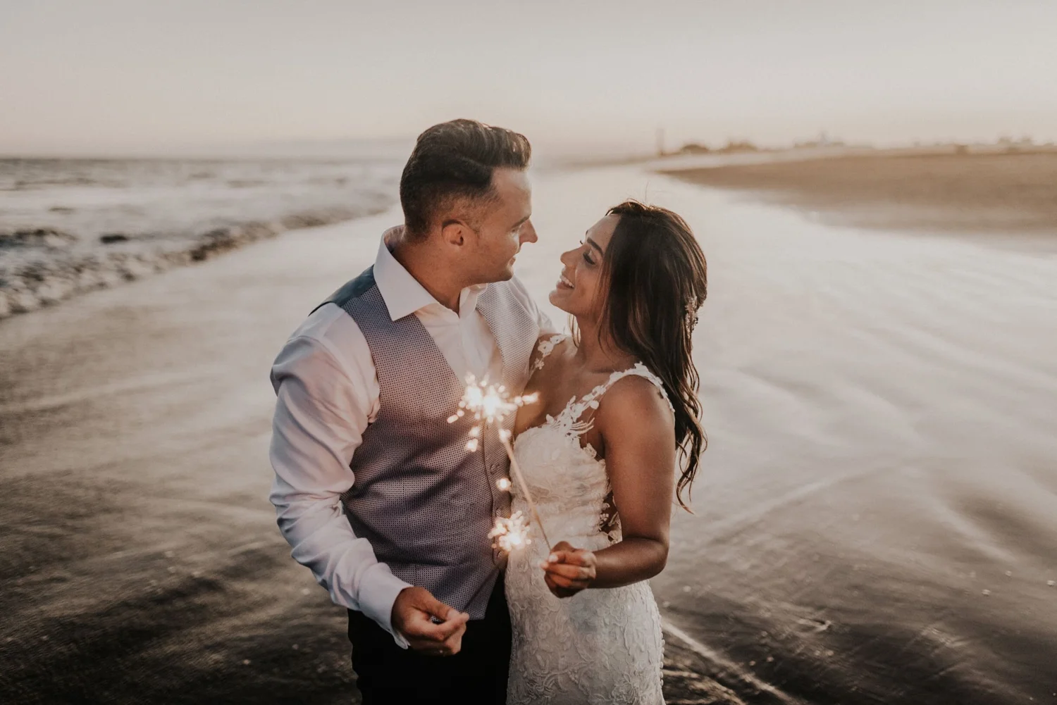 A newlywed couple smiling and holding sparklers on the beach at sunset.