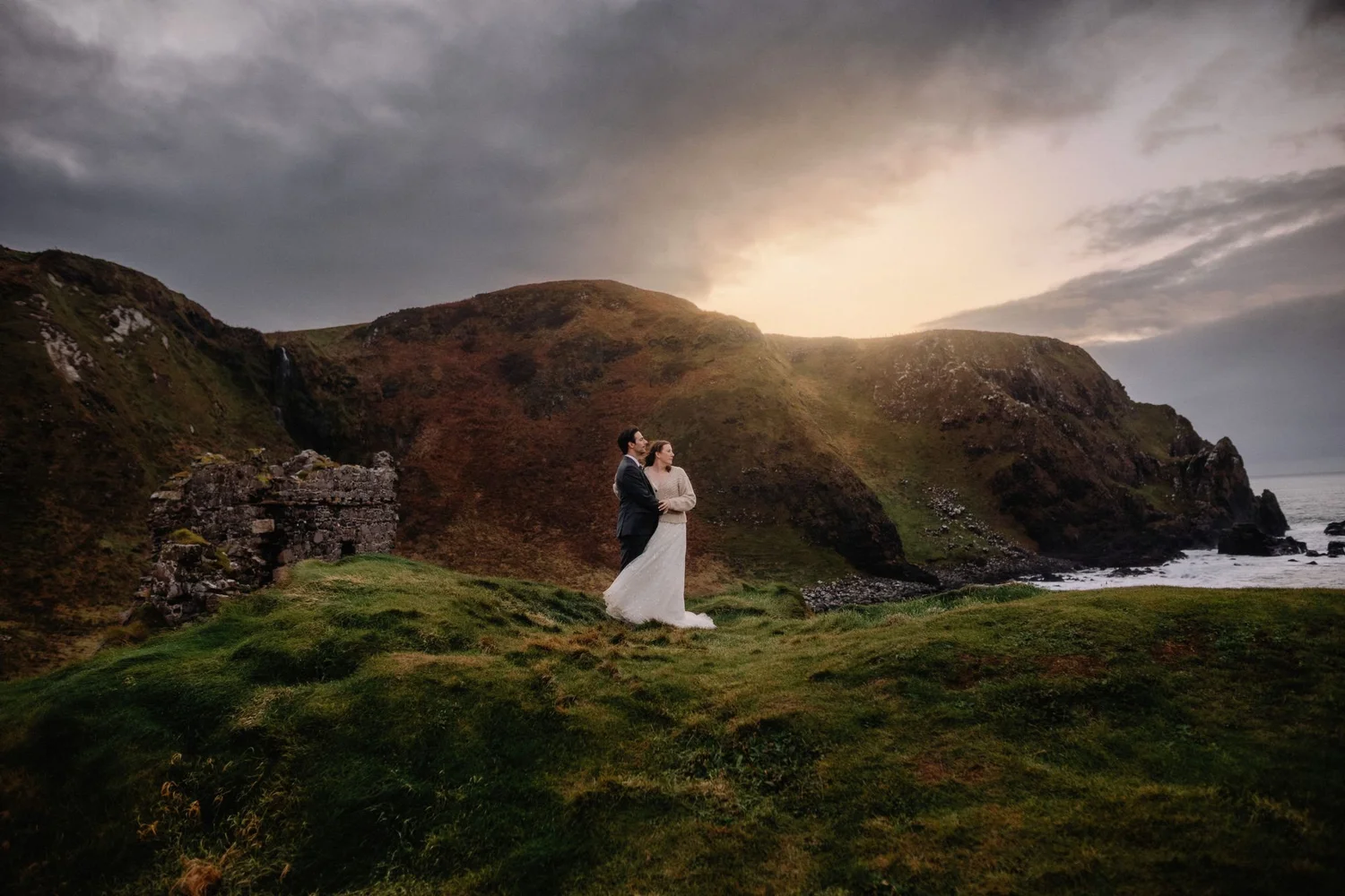 A bride and groom standing together on a grassy cliffside overlooking the ocean with mountainous terrain in the background during sunset or sunrise.