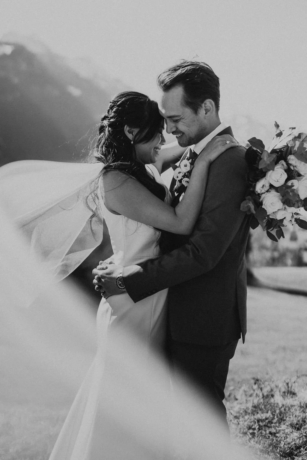 A black-and-white photo of a bride and groom embracing outdoors, smiling and touching foreheads, with the bride holding a bouquet of flowers.