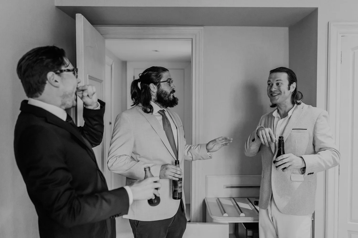 Three men in suits and one in a tuxedo, holding beer bottles, engaged in conversation in a room with light-colored walls and a doorway.