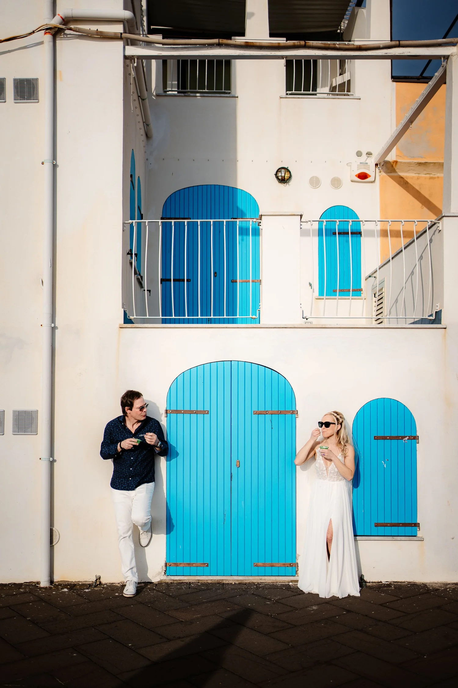 A man and a woman standing against a white wall with blue doors and shutters, with the man talking and the woman adjusting her sunglasses.