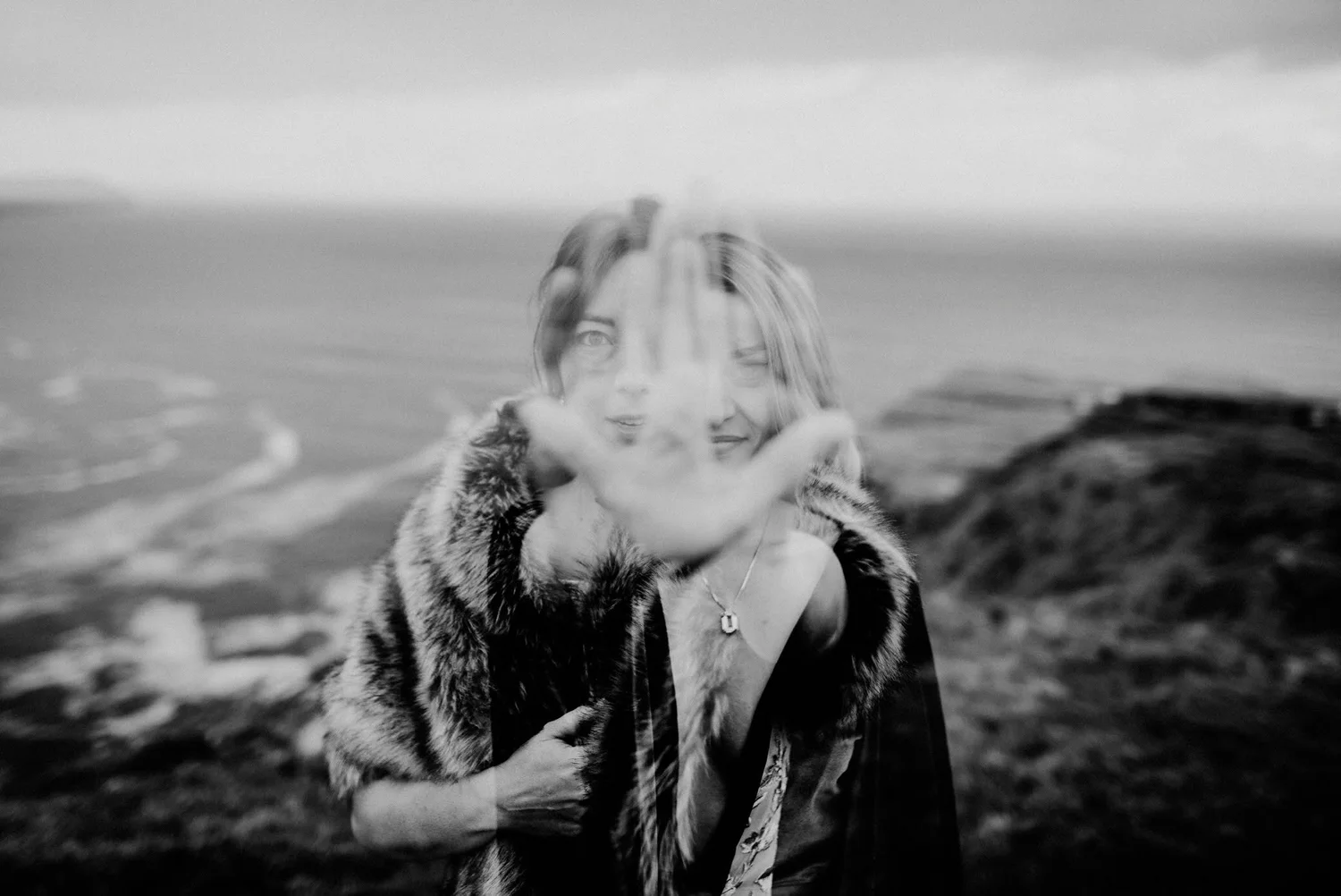 A woman in a faux fur coat holds her hand up to her face, partially covering her expression, with a landscape background in black and white.