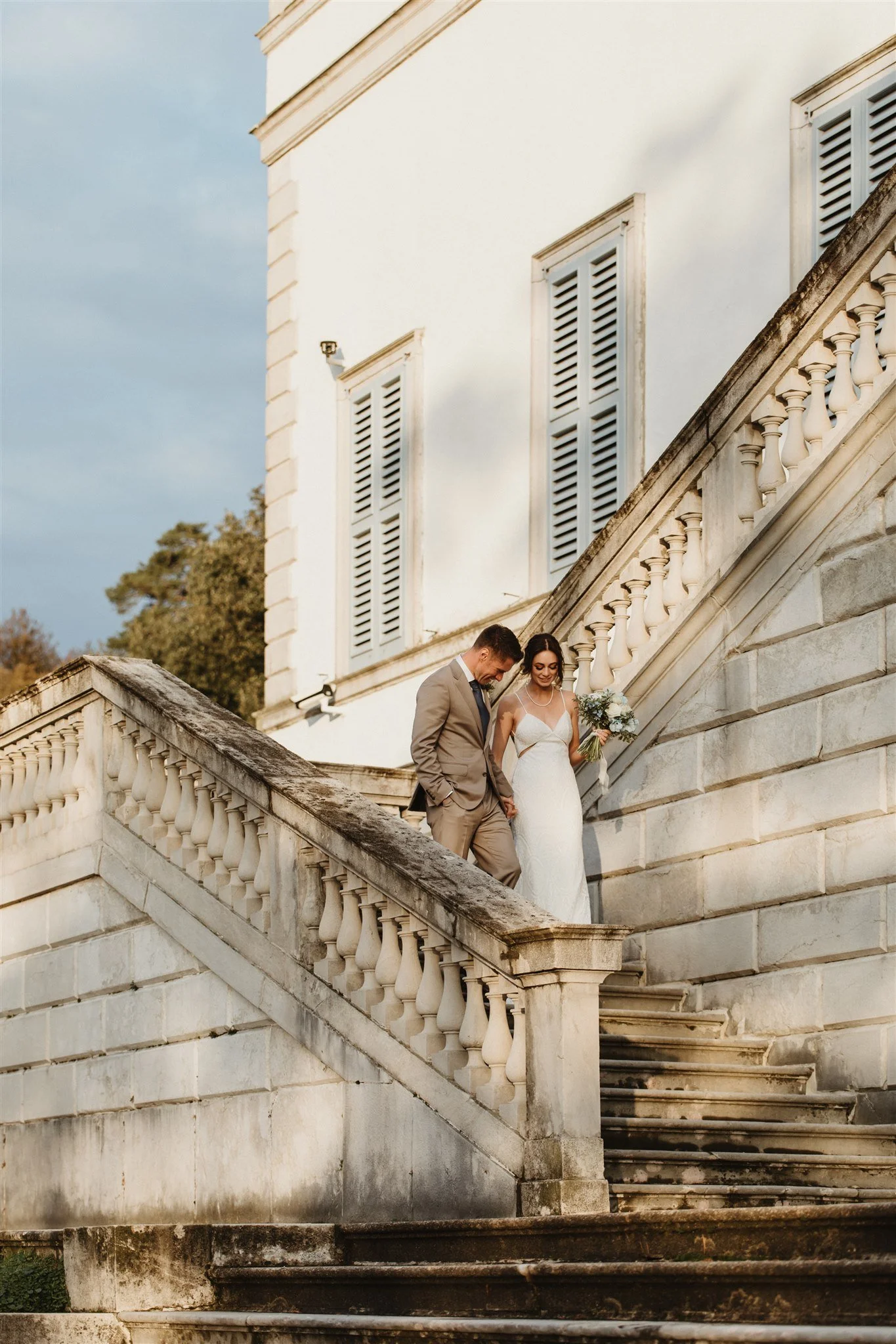 A bride and groom dressed in wedding attire walking down an outdoor stone staircase with a white building in the background.