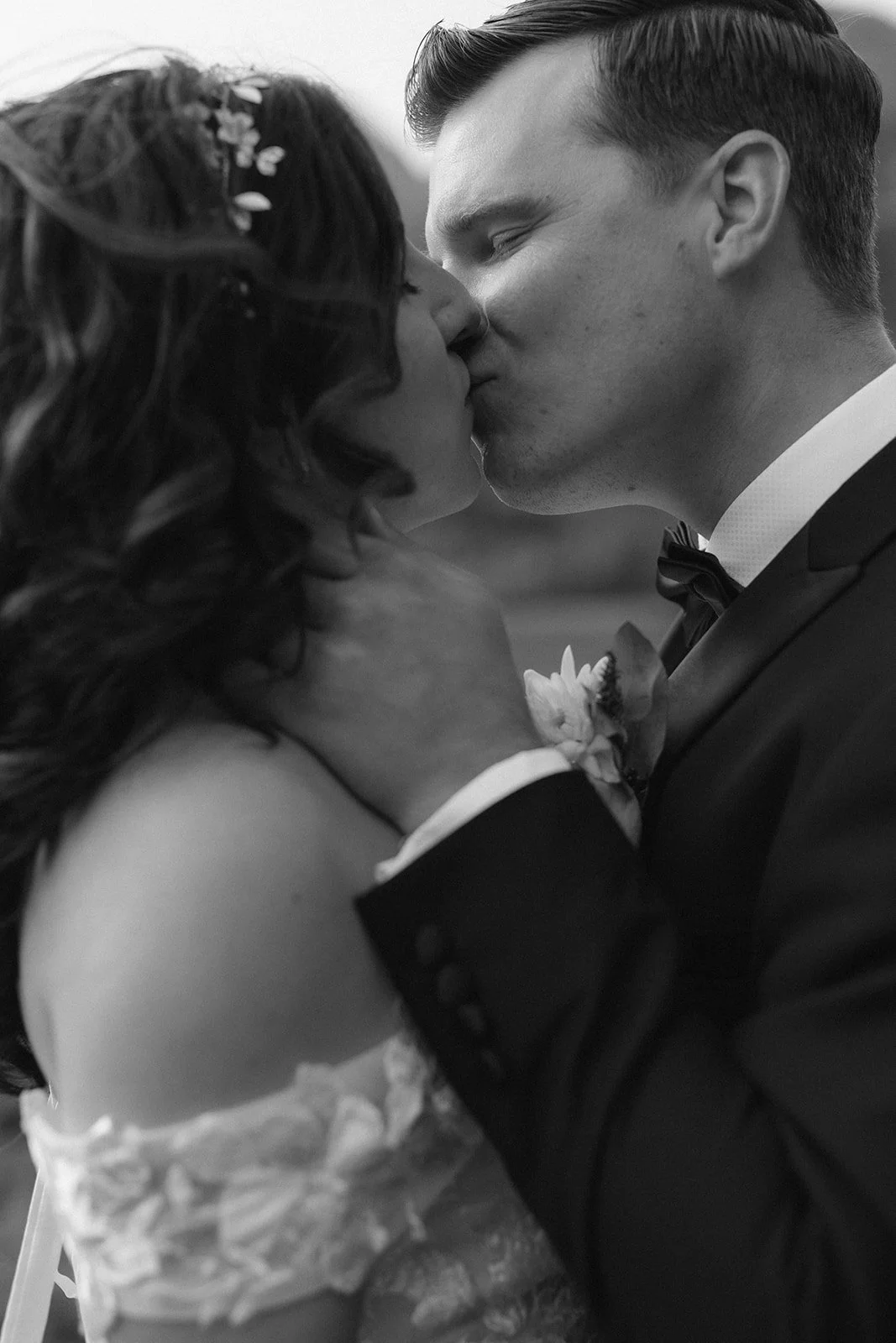 Black and white photo of a couple kissing, the woman has long wavy hair with flowers, and the man is wearing a tuxedo with a bow tie and a floral boutonniere.