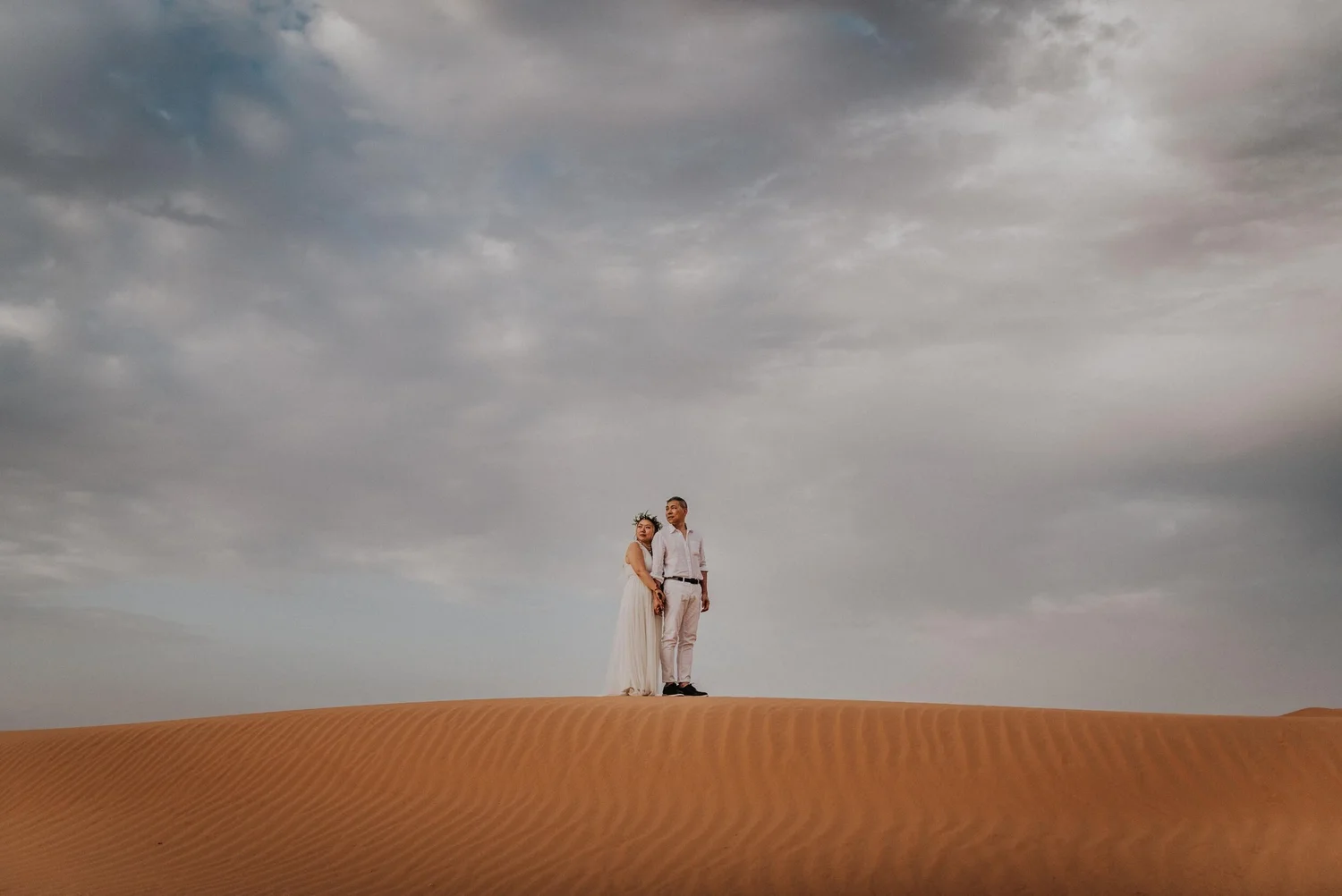 A couple stands close together on a sandy desert landscape under a cloudy sky, holding hands and looking serenely into the distance.