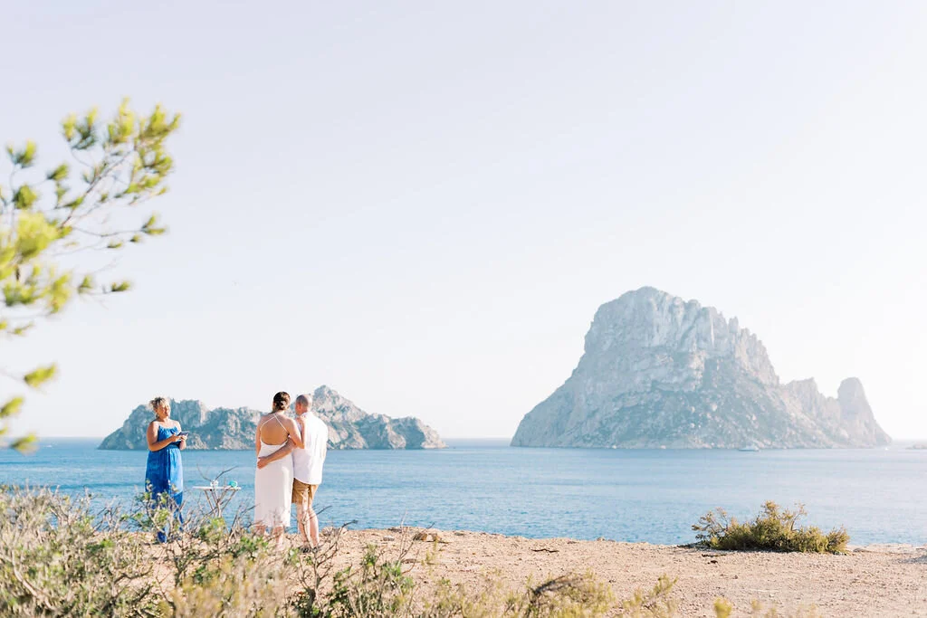 A couple getting married on a beach, with a woman in a blue dress officiating, and large rocky islands in the background.