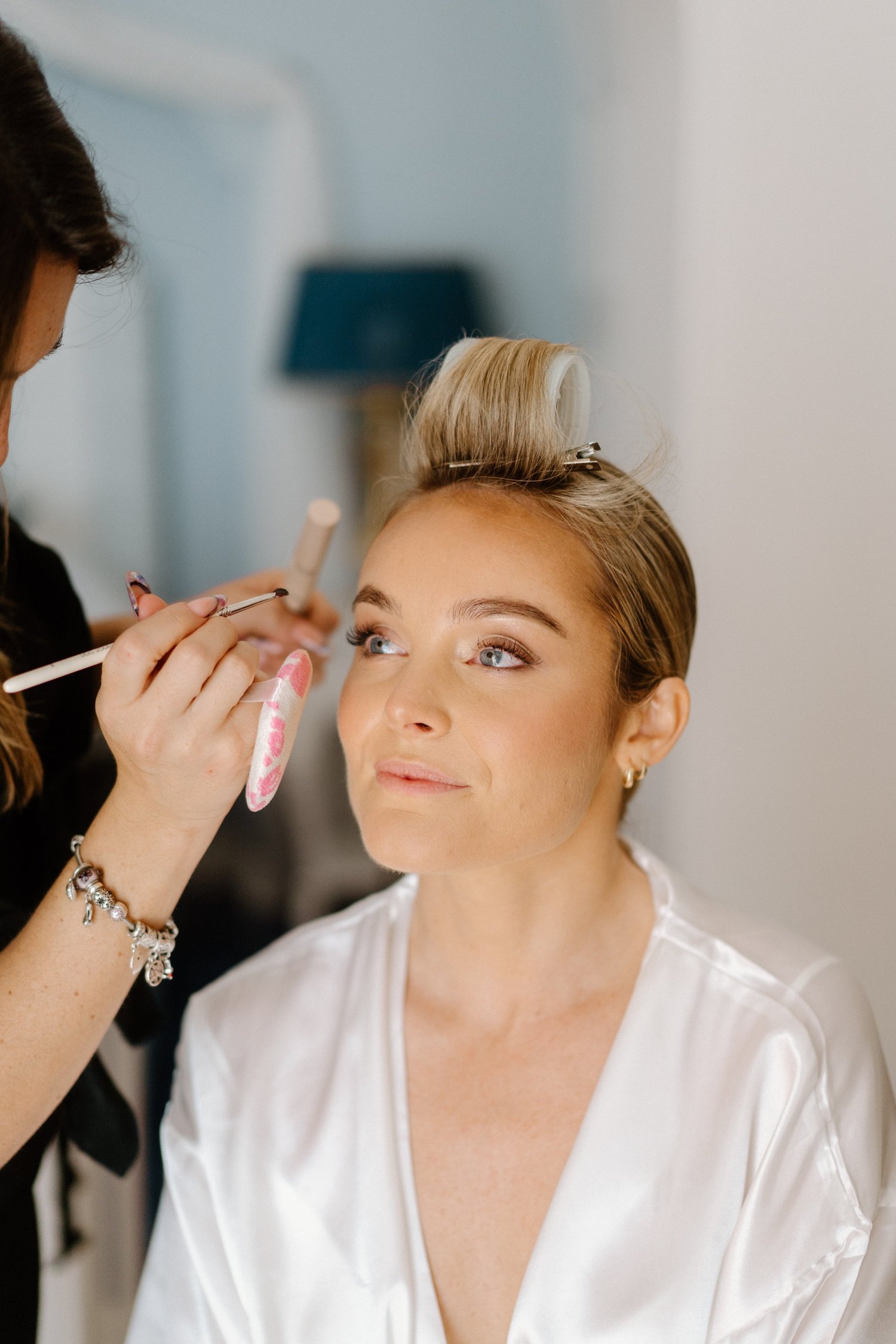 A woman with blonde hair is getting her makeup done by a makeup artist. She is wearing a white satin robe and has a curlers in her hair. The woman is sitting and looking slightly upwards.