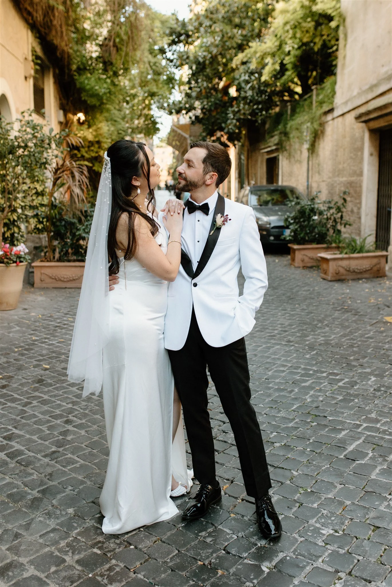 A bride and groom stand close together on a cobblestone street lined with potted plants and old buildings, sharing an intimate moment during their wedding day.