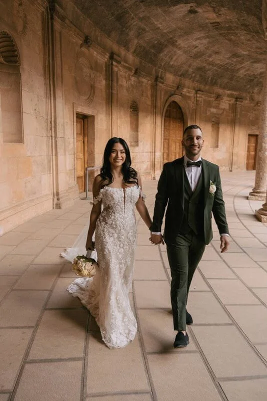 A bride and groom walking hand in hand in a historic architectural setting with stone walls and columns, both dressed in wedding attire, smiling and looking happy.