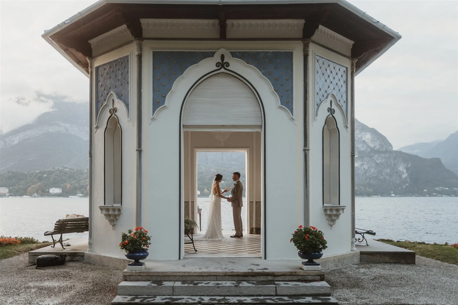 A bride and groom stand facing each other under a decorative pavilion near a body of water, with mountains in the background. The bride is in a white gown and the groom in a tan suit, holding hands and swearing vows.
