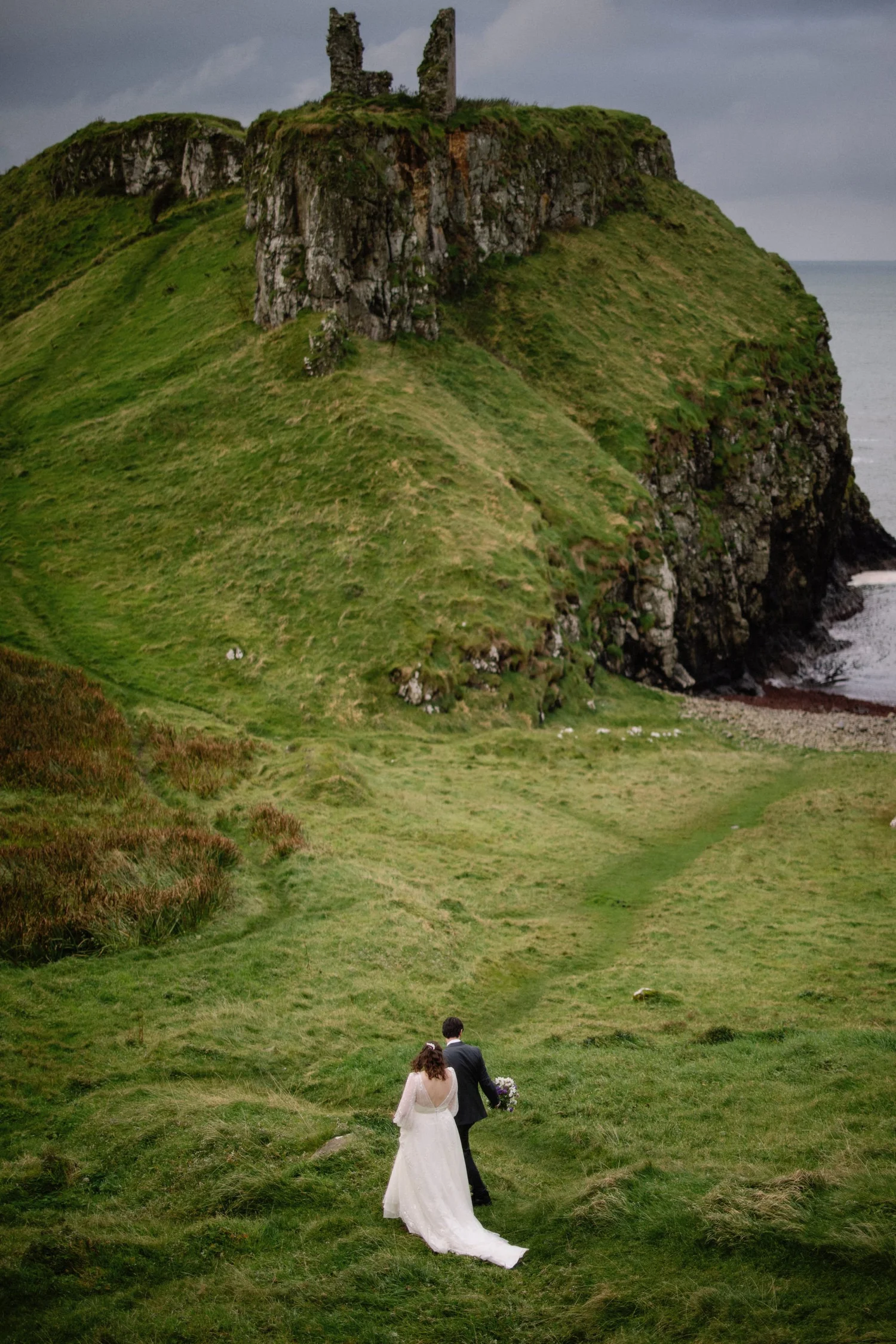 A newlywed couple walking on a grassy hillside near a rocky cliff and the ocean, with the remains of an old stone building on top of the hill.