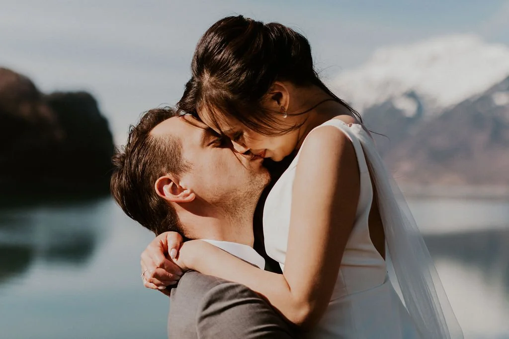 A couple embraces and kisses outdoors near a lake with mountains in the background.