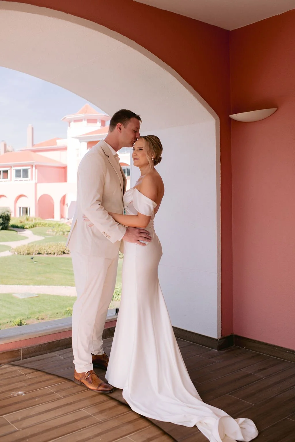 A couple in wedding attire sharing a moment indoors, with an outdoor scene of a large pink building and green lawn visible through an arched window behind them.