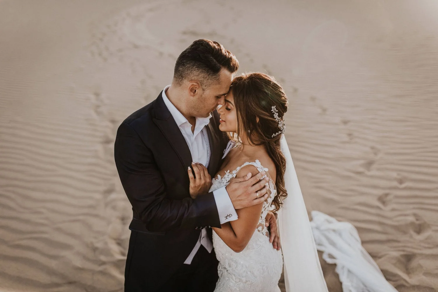 A bride and groom embrace on the beach, their foreheads touching, with the ocean in the background.