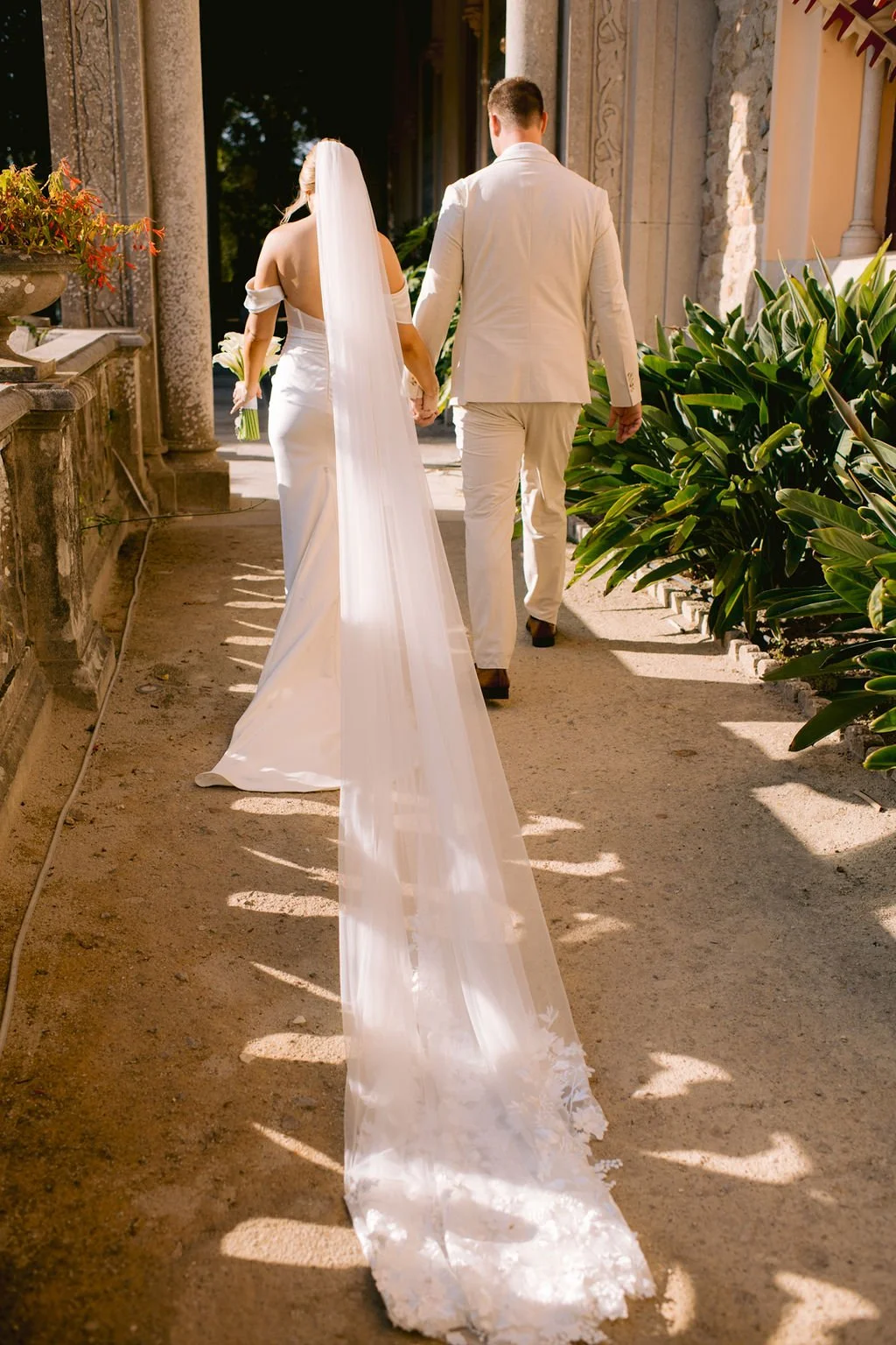 Bride and groom walking hand in hand outside a church or venue, with the bride wearing a white wedding gown and veil, holding a bouquet of flowers, and the groom dressed in a light-colored suit.